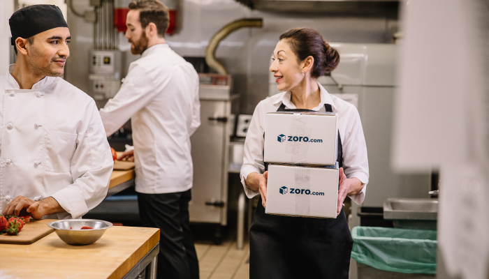 Image of a worker carrying 2 boxes labelled "Zoro.com" on the sides through a large commercial kitchen area.