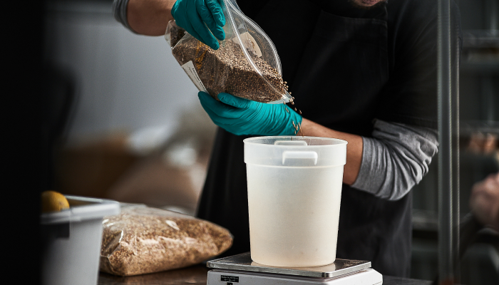 Image of person with gloves on, pouring grains from a clear plastic bag into a round plastic container sitting on a weight scale.